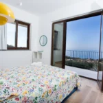Camera matrimoniale con vista mare, lampade gialle e biancheria floreale – Villa Giardino degli Ulivi