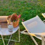 Dettaglio di tavolino da esterno con bicchieri, bottiglia d’acqua, giornali e fiori freschi in un giardino con ulivi a Capo d'Orlando.