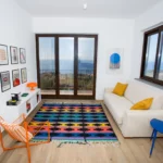 Zona relax con tappeto etnico, sedia arancione, divano bianco e vista sul mare – Villa Giardino degli Ulivi, Sicilia