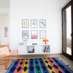 Angolo lettura con tappeto etnico, sedia arancione e stampe grafiche su parete bianca – Villa Giardino degli Ulivi, Sicilia