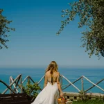 Donna con abito bianco e cestino di frutta cammina sul prato di Villa Giardino degli Ulivi, con vista mare e l’isola di Alicudi all’orizzonte.