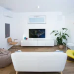 Zona living moderna con divano bianco, poltrona gialla e televisore, decorata con piante e dettagli di design – Villa Giardino degli Ulivi, Sicilia
